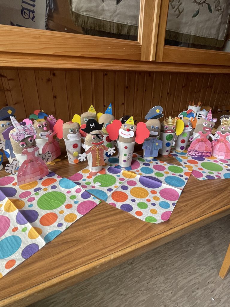 Eine farbenfrohe Reihe von kreativ verkleideten Blumenzwiebeln und Töpfen – gestaltet als Piraten, Clowns, Prinzessinnen und Polizisten – steht auf bunten, gepunkteten Servietten auf einem Holztisch als Ergebnis des „Beet Buddies“-Bastelworkshops.