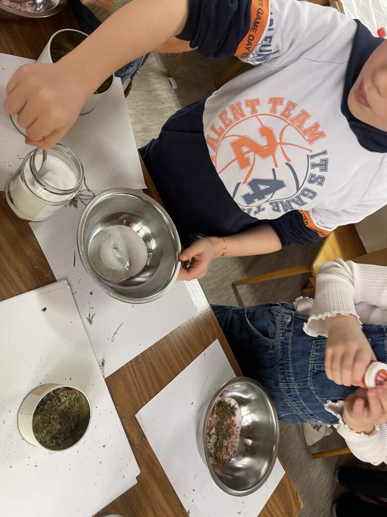 Hohe Draufsicht auf einen Holztisch, an dem zwei Kinder konzentriert an der Herstellung von eigenem Pizzasalz arbeiten. Das obere Kind, in einem weißen "TALENT TEAM"-Langarmshirt mit orange-blauen Akzenten, löffelt mit einem Löffel Salz aus einem Glas in eine Metallschüssel, die bereits einen Kräuter-Mix enthält. Das untere Kind, in einer Jeans-Latzhose und einem weißen Rüschen-Shirt, mischt mit bloßen Händen einen anderen Kräuter- und Salz-Mix in einer separaten Metallschüssel. Der Tisch ist übersät mit offenen Metalldosen voller getrockneter Kräuter (wie Thymian und Rosmarin), Gläsern mit Salz, Rührschüsseln, leeren kleinen Glasfläschchen und goldenen Deckeln. Die Szene ist hell und die Holzstruktur des Tisches und der Wand im Hintergrund ist sichtbar.
