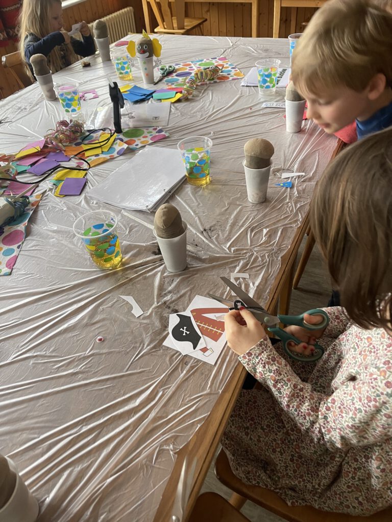 Kinder sitzen an einem langen, mit Folie gedeckten Tisch und basteln im Rahmen des „Beet Buddies“-Workshops „Verkleidungskünstler im Garten“. Ein Mädchen schneidet mit einer Schere Papier-Accessoires wie einen Piratenhut aus, während auf dem Tisch Töpfe mit Blumenzwiebeln und bunte Bastelmaterialien liegen.