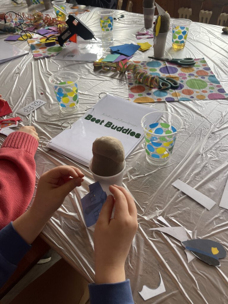 Ein Kind bastelt an einem Tisch voller Bastelmaterialien, Luftschlangen und bunter Papiere an einer verkleideten Blumenzwiebel im Becher; auf dem Tisch liegt ein Ordner mit der Aufschrift „Kinder- und Jugendgruppe Beet Buddies“
