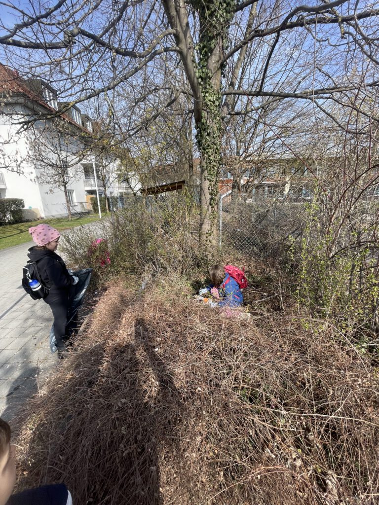 Kinder nehmen an einer Ramadama-Aktion der Beet Buddies teil: Ein Kind mit rosa Mütze hält am Wegrand einen Müllsack bereit, während ein weiteres Kind mit rotem Rucksack in den Büschen nach Müll sucht.