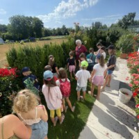 Eine Gruppe von Kindern erkundet beim Sommerabschluss der Beet Buddies den sonnigen Garten des Vorstandsvorsitzenden, der von üppigen Blumen und grünen Hecken umgeben ist.