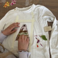 Ein Kind gestaltet im Rahmen des Beet-Buddies-Workshops ein weißes Langarmshirt mit Naturfarben, indem es ein mit Klebeband fixiertes Motiv auf dem Stoff anreibt, während auf dem Holztisch ringsherum bunte Blütenblätter und grüne Blätter verstreut liegen.
