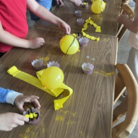 Mehrere Kinder stehen an einem langen Holztisch und simulieren mit gelben Ballons, kleinen Förmchen als Blüten und gelbem Blütenstaub spielerisch die Bestäubung durch Bienen.