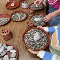 Kinder der Beet Buddies basteln an einem Holztisch eigene Bienentränken aus Pflanzuntersetzern, kleinen Kieselsteinen und größeren Steinen als sichere Wasserstellen für Bienen und Insekten.
