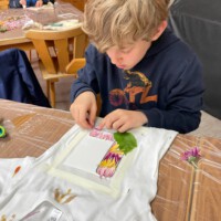 Ein blonder Junge in einem dunkelblauen Hoodie platziert konzentriert bunte Blütenblätter und grüne Blätter auf einem weißen T-Shirt, um im Rahmen des Beet-Buddies-Workshops ein natürliches Muster zu gestalten.