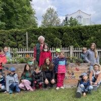 Eine große Gruppe von „Beet Buddies“-Kindern und Betreuern posiert fröhlich im Garten vor und hinter zwei bepflanzten Holz-Hochbeeten, die mit bunten Handabdrücken und Schriftzügen verziert sind.