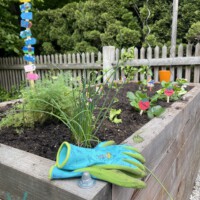 Ein Paar blau-grüne Gartenhandschuhe liegt auf dem Rand eines hölzernen Hochbeets, in dem Kräuter und Erdbeeren mit kleinen Schildern wachsen; im Hintergrund sind ein Gartenzaun und ein bunter Wegweiser zu sehen.