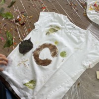 Ein selbst gestaltetes weißes T-Shirt mit natürlichen Pflanzenfarben sowie Abdrücken von Blättern und Blüten liegt auf einem mit Folie ausgelegten Tisch, umgeben von verstreuten bunten Blütenblättern, grünen Blättern und einer Musterschablone im Rahmen des Beet-Buddies-Workshops.